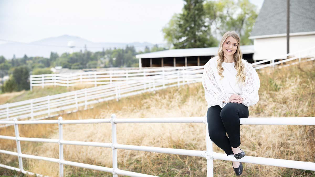 A woman sits on a white fence with a white barn in the background.