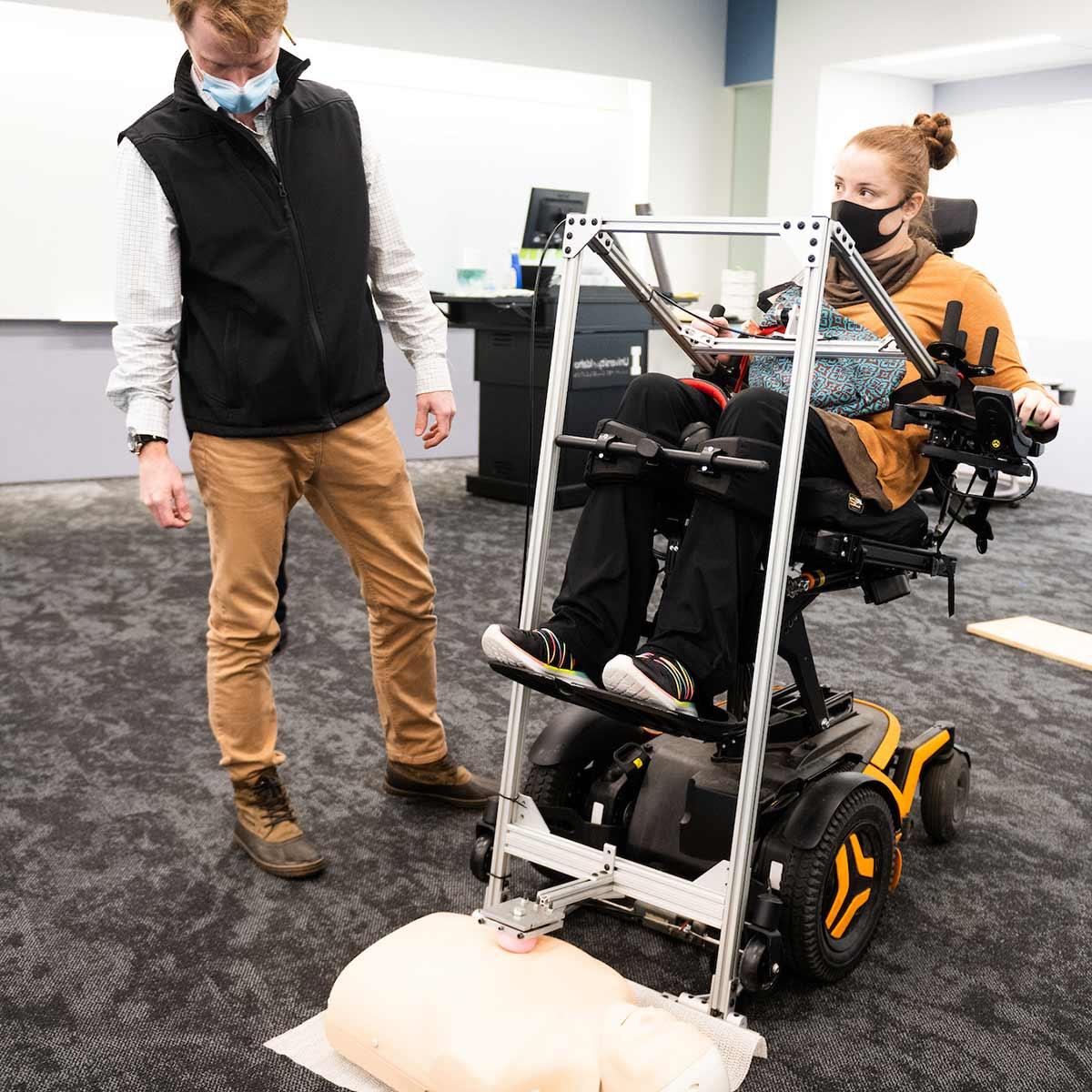 Engineering students help a student in a wheelchair use the assistive CPR device, which uses a lever-action to augments force and achieve necessary chest compression.